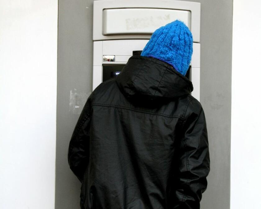 Man met zwarte jas, blauwe muts en jeansbroek neemt portefeuille uit achterzak. Hij staat voor een geldautomaat. In blauwe letters op gele achtergrond staat 'cash' boven de automaat.