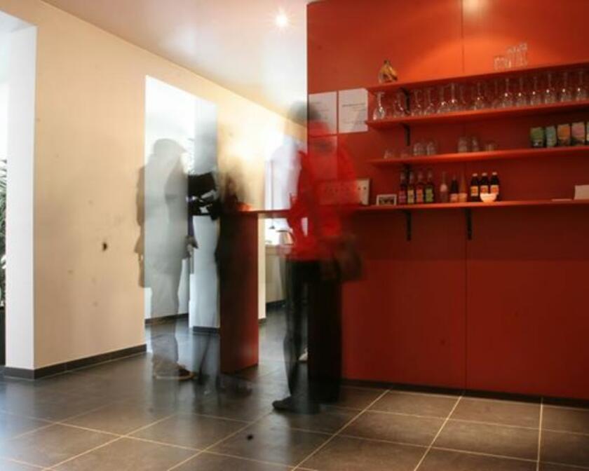 Rode muur met rekken met glazen en flesjes, grijze vloer met grote tegels, wazige mensen.