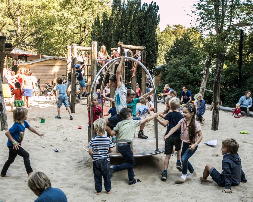 Eine große Gruppe von Kindern spielt auf verschiedenen Holz- und Matallspielzeugen. Der Boden besteht aus Sand. Es ist ein Sommertag.