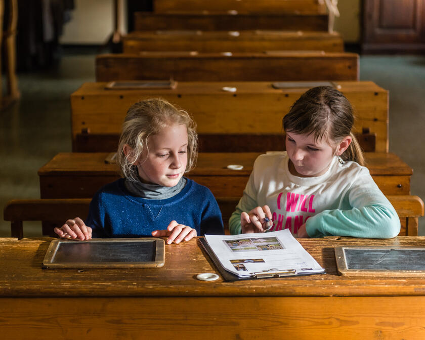 Twee schoolmeisjes in een klaslokaal aan een oude houten schoolbank met leitjes voor zich en een invulblad in het Museum De school van Toen; een rij lege bankjes is te zien achter de eerste bank.