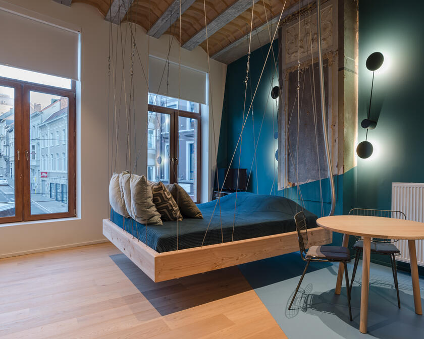 kamer met tweepersoonsbed dat aan het plafond hangt, blauwe en witte elementen, een tafel met 2 stoelen