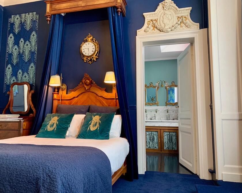 blauwe slaapkamer met tweepersoonsbed, blauwe kussens met kever op, blauwe bedgordijnen, deur naar badkamer