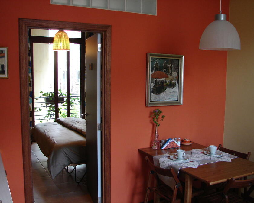 kamer met houten tafel en stoelen, rode muur, deur die uitkomt op slaapkamer, schilderij aan de muur