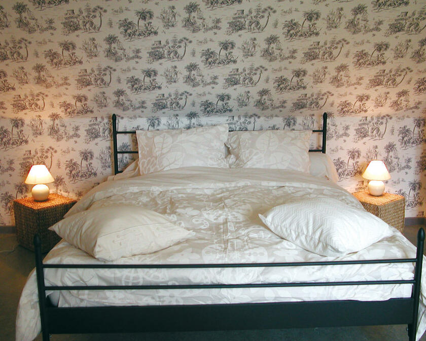 kamer met tweepersoonsbed, 2 nachtkastjes met 2 lampen, wit behang met palmbomen en mensen