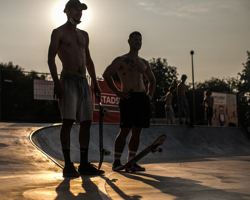Twee skaters staan aan de rand van het skatepark, een met een skateboard in de hand en de andere met een skateboard aan de voet