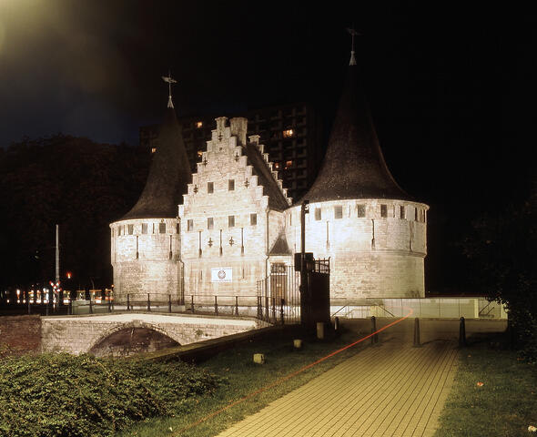 Verlicht grijs gebouw met twee ronde torens langs het water bij avond