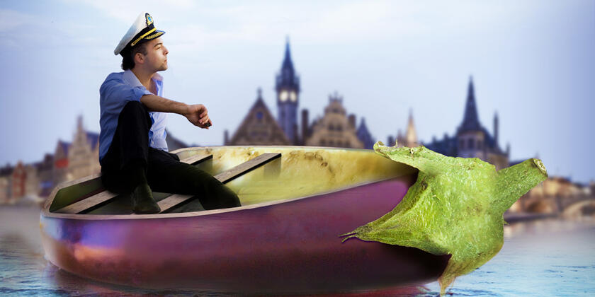 Un chico con gorra de patrón sentado en un barco en forma de berenjena, con el Graslei de fondo