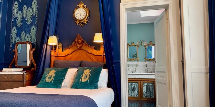 blauwe slaapkamer met tweepersoonsbed, blauwe kussens met kever op, blauwe bedgordijnen, deur naar badkamer