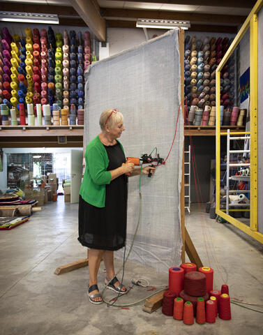 een vrouw die rode draad vastmaakt aan een grijs doek, tegen de muur rekken vol met touw in verschillende kleuren
