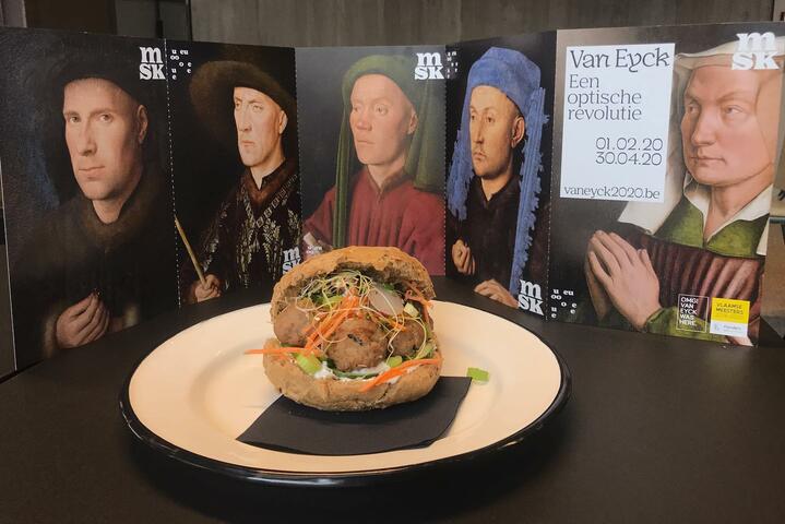 een bord met een broodje met gehaktballen en groenten, hierachter folders van van eyck