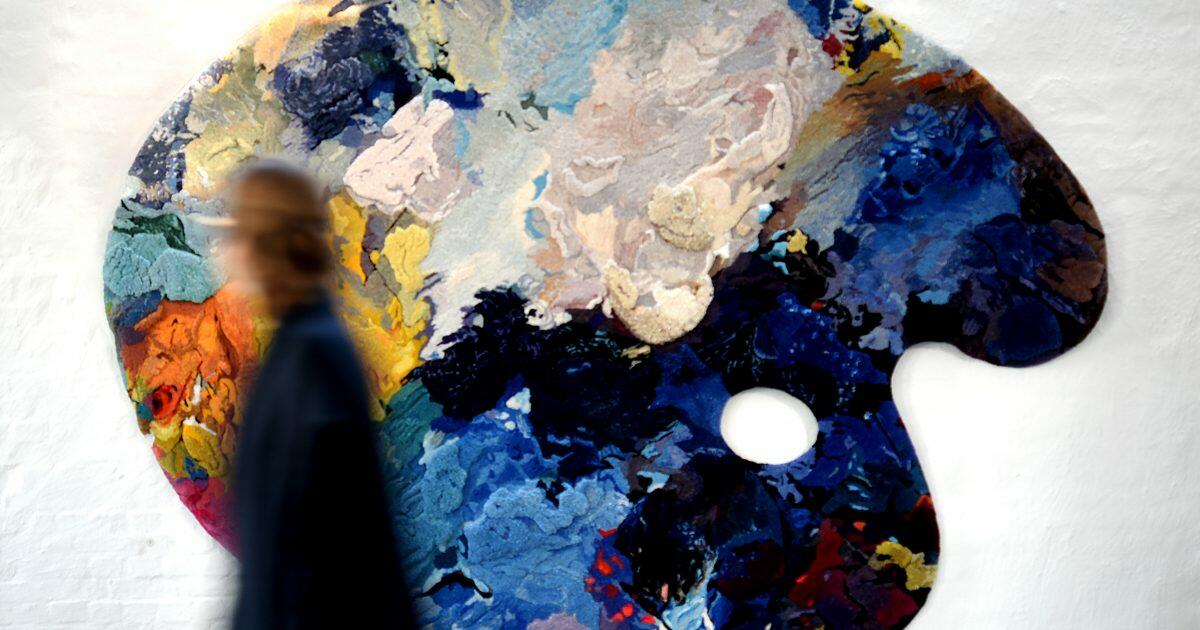 groot schilderspalet met verschillende kleuren verf op tegen een witte muur, iemand die er voorbij wandelt