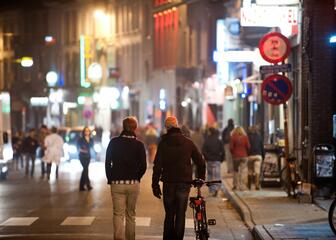 Twee jongeren (één met de fiets aan de hand) stappen weg van de fotograaf, richting een drukke straat met cafés en veel mensen; 's nachts.
