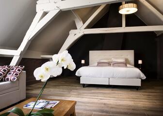 Slaapkamer onder het dak. Het beige bed en gebinte contrasteren met de donkerbruine muur.
