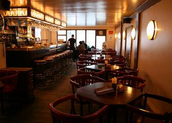 Binnenzicht met kleine vierkante tafels en brede armstoelen en zicht op de bar met hoge barkrukken.