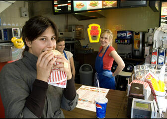 Zicht op toog met twee dames in blauwe schort en één dame op de voorgrond die een hap neemt van een hamburger.