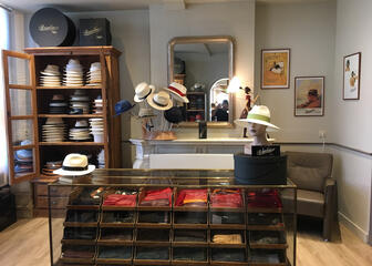 Het interieur van een hoedenwinkel. In het midden is er een glazen kast en er liggen verschillende sjaals en handschoenen. 