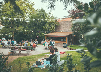 Mensen zitten op het gras of aan een tafel in een versierd tuintje met vlagjes en lichtjes. Ze genieten van een hapje en een drankje.