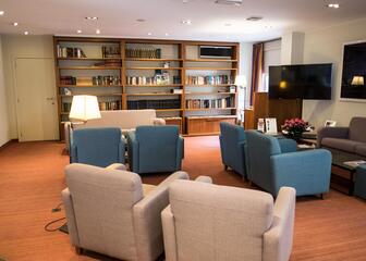 Ontvangsthal met beige, blauwe en grijze zetels. In de kamer zie je eveneens een televisie en grote boekenwand. 