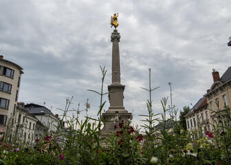 monument met gouden beeldje vanboven, gebouwen langs beide kanten, veel bloemen ervoor
