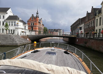 het voordek van een boot op het water, voor een brug met gebouwen langs de zijkanten