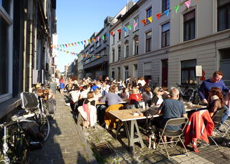 Straatfeest met mensen aan lange tafels, gekleurde vlaggetjes zijn gespannen tussen de huizen boven de straat.
