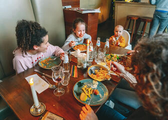 Eettafel met één baby en twee jonge kinderen met ouders. De tafel is gevuld met eten (spaghetti, frietjes…)
