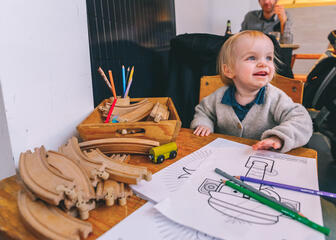 Blonde kleuter in kinderstoel met tekeningen en kleurpotloden aan tafel in Lekker Gec.