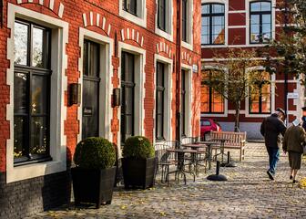 Tafeltjes, stoelen en plantenbakken voor rood huis in het Oud Begijnhof, kasseistraat, met een ouder koppel op wandel.