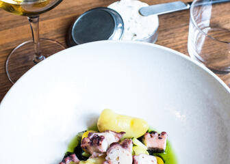Octopushapje, aardappel, groene emulsie op wit bord, broodjes met tapenade, glas witte wijn.