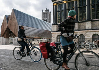 Twee fietsers met de achtergrond de stadshal, de toren van de Sint-Niklaaskerk en een deel van de gevel van het stadhuis. 