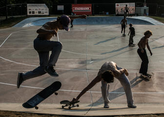 Een skater doet een trick met zijn skateboard, terwijl een andere skater zijn skateboard opraapt. In de achtergrond zijn nog vijf andere skaters te zien.