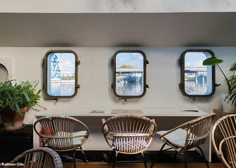 binnenkant boot met tafel en stoelen, planten, 3 ramen die uitkijken op het water