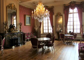 salon met antieke tafels en stoelen, kroonluchter, spiegel, bustes, schilderij, rode gordijnen aan de ramen