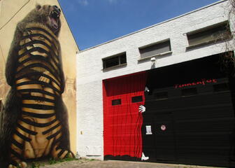 wit gebouw met zwarte garagepoort met in het rood Tinnenpot en een persoon achter een rood gordijn op,  gebouw ernaast met street art van een beer die een leeuw vasthoudt