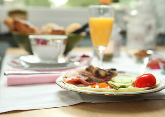 Detailaufnahme des Frühstückstisches. Das reichhaltige Frühstück umfasst frisch gepressten Fruchtsaft, Kaffee, ein Spiegelei, einen Streifen Speck, ein Stück Tomate, Gurke und Pilze