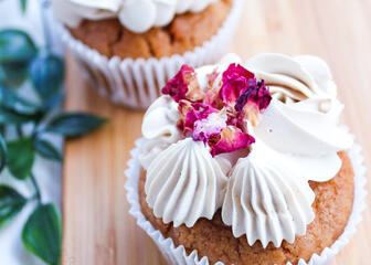 2 cupcakes with white frosting and flower petals on a wooden board 