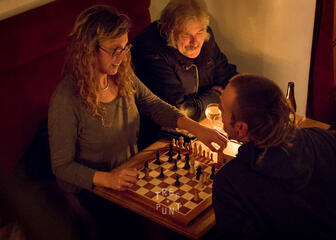 een vrouw en 2 mannen zitten aan een tafel met een schaakbord tussen hen
