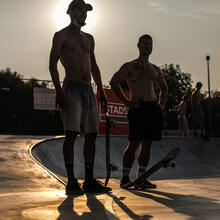 Twee skaters staan aan de rand van het skatepark, een met een skateboard in de hand en de andere met een skateboard aan de voet