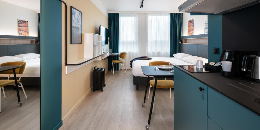 vue d’une chambre à coucher avec cuisine moderne, coin repas, lit noir et, à gauche, un bureau suspendu blanc avec une chaise jaune foncé 