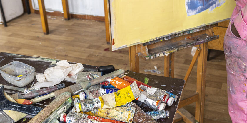 Paint brushes and paint on a table, a colourfully painted canvas in the background.