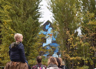 Een groep mensen met een kleine jongen op de schouders van een ouder op de voorgrond. Op de achtergrond is tussen de bomen door een graffiti werk zichtbaar van een blauw figuur.