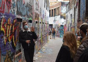 Een man volledig in het zwart gekleed en met mondmasker leunt tegen de muur terwijl hij uitleg geeft in het graffitistraatje.
