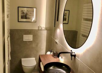 baño con lavabo marrón, espejo redondo iluminado e inodoro
